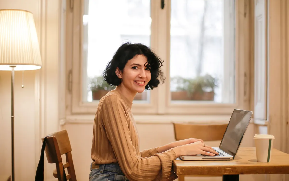 A woman smiling over her laptop at a café table by the window, a coffee beside her